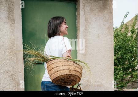 Seitenansicht einer fröhlichen jungen hispanischen Farmerin mit dunklem Haar in legerer Kleidung, die handgefertigten Korbkorb mit Esparto-Gras hält und lächelt in c Stockfoto