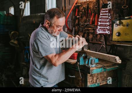 Fokussierter reifer männlicher Holzarbeiter, der Tischler für die Planung von Holzplanken verwendet, während er an einer schäbigen Werkbank in der Schreinerei arbeitet Stockfoto