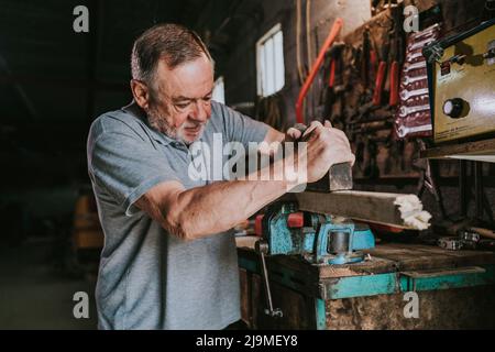 Fokussierter reifer männlicher Holzarbeiter, der Tischler für die Planung von Holzplanken verwendet, während er an einer schäbigen Werkbank in der Schreinerei arbeitet Stockfoto