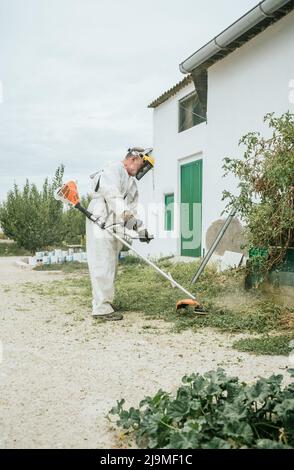Ganzkörper-Seitenansicht des gealterten, nicht erkennbaren männlichen Bauern in weißer Overall-Kleidung und Schutzmaske, die Gras mit Unkrautmesser schneidet, während sie in g arbeitet Stockfoto