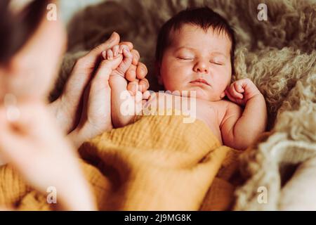 Hoher Winkel von unerkennbarer junger Mutter und Vater mit dunklem Haar, die Hand des entzückenden Neugeborenen haltend, das bei Tageslicht auf einem weichen Bett liegt und schläft Stockfoto