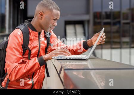 Seitenansicht eines fokussierten jungen afroamerikanischen Studenten in stilvollem Outfit und Rucksack, der auf der Straße steht und während der Vorbereitung auf die Prüfung den Computer benutzt Stockfoto
