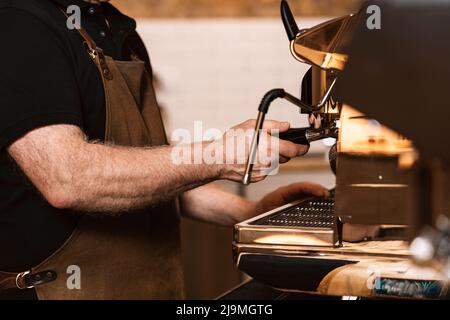 Seitenansicht des älteren Barista in der Schürze, der den Portafilter in eine moderne professionelle Kaffeemaschine legt, während er im Café Espresso brüht Stockfoto