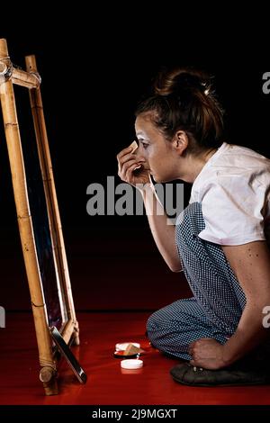 Junge professionelle weibliche Pantomime in legerer Kleidung, die ein Fundament auf dem Gesicht aufträgt und im Spiegel sitzt, während sie auf dem Boden in der Ankleidekabine sitzt Stockfoto