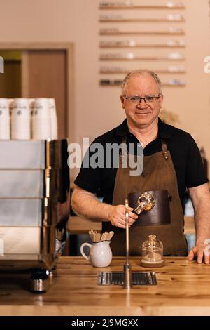Positiver reifer Barista mit Schürze und Brille, lächelnd und mit Blick auf die Kamera, während er an der Holztheke in der Nähe der modernen Kaffeemaschine mit porta stand Stockfoto