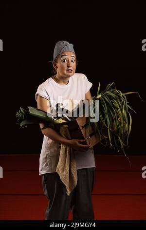 Überrascht junge Clownin schmutziger Kleidung und Schürze mit lustigen Make-up Blick auf die Kamera, während sie Korb mit frischem Lauch und Stahl Schöpflöffel Agai Stockfoto