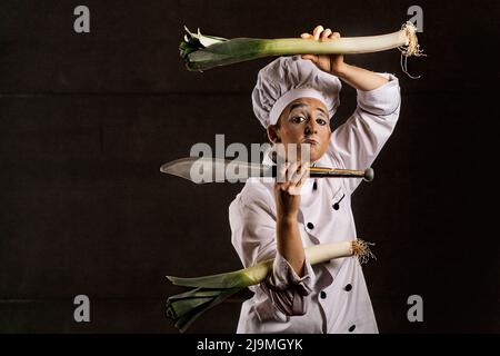 Verärgert weibliche Pantomime Künstlerin mit Make-up in Koch Kostüm und Hut machen trauriges Gesicht und Blick auf die Kamera, während sie frische Lauch und scharfe Messer gegen b Stockfoto