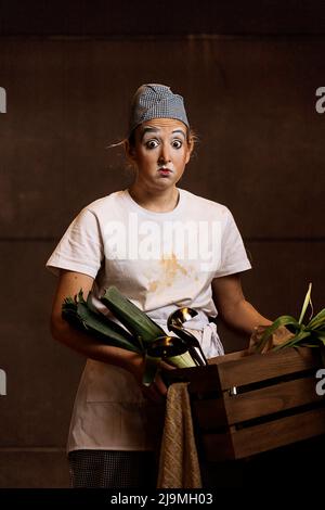 Überrascht junge Clownin schmutziger Kleidung und Schürze mit lustigen Make-up Blick auf die Kamera, während sie Korb mit frischem Lauch und Stahl Schöpflöffel Agai Stockfoto