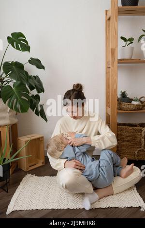 Junge Mutter stillt entzückendes kleines Kind mit blonden Haaren, das auf dem Boden in einer modernen Wohnung bei Tageslicht sitzt Stockfoto