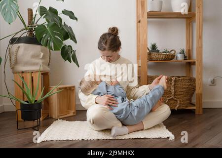 Junge Mutter stillt entzückendes kleines Kind mit blonden Haaren, das auf dem Boden in einer modernen Wohnung bei Tageslicht sitzt Stockfoto