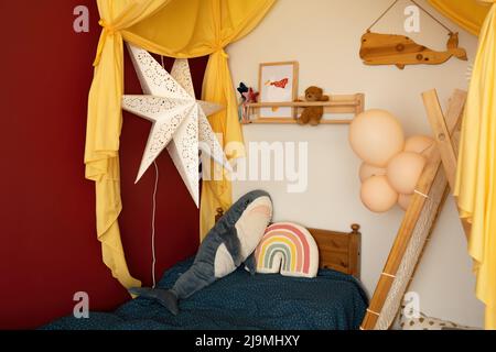 Gelbes Baldachin über einem komfortablen Bett, das mit einem weichen Hai und regenbogenförmigen Kissen in einem gemütlichen Kinderzimmer dekoriert ist Stockfoto