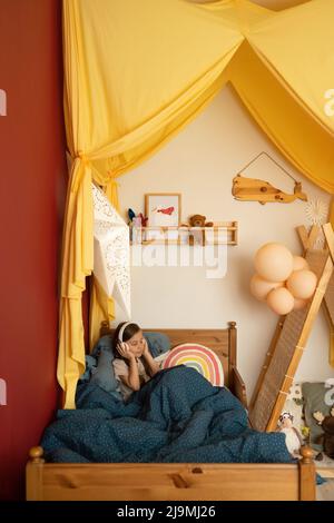 Entzückendes kleines Kind mit langen dunklen Haaren, die unter einer warmen Decke auf dem Bett liegen und in einem gemütlichen, mit Baldachin dekorierten Schlafzimmer Musik über Kopfhörer hören Stockfoto