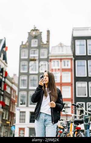 Niedriger Winkel der ernsthaften jungen ethnischen weiblichen Touristen mit langen dunklen Haaren in trendigen Outfit Aufnahme Sprachnachricht über Handy, während sich dagegen Stockfoto