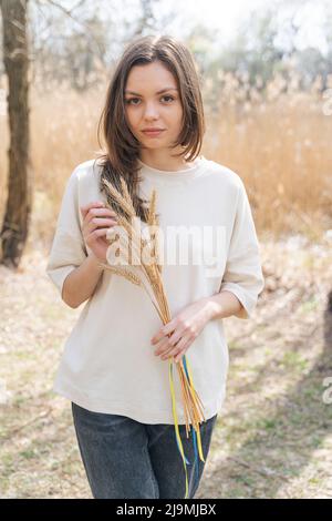 Ernst junge Frau mit langen dunklen Haaren in lässigen Kleidern lächeln und Blick auf die Kamera, während in der Natur mit Haufen Weizenspikelets gebunden w stehen Stockfoto