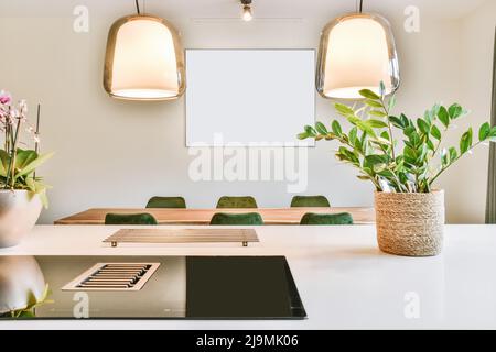 Ein leeres Bild hängt an der Wand über dem Tisch mit Stühlen hinter Lampen und einer Theke mit Topfpflanzen in der modernen Küche tagsüber Stockfoto