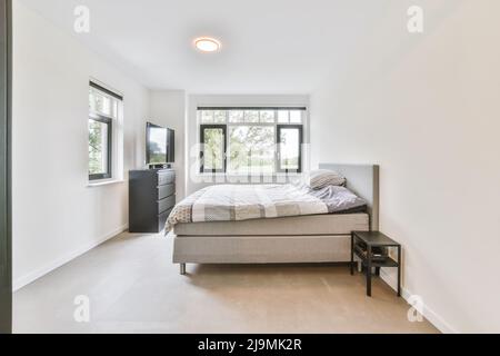 Komfortables Bett mit Federdecke und Kissen in der Nähe des Fensters und schwarzem Schrank mit fernseher im hellen Schlafzimmer zu Hause Stockfoto