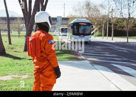 Seitenansicht einer nicht erkennbaren Person im leuchtend orangefarbenen Astronauten-Anzug und Helm, die an sonnigen Tagen in der Stadt mit modernen Bussen am Straßenrand steht Stockfoto