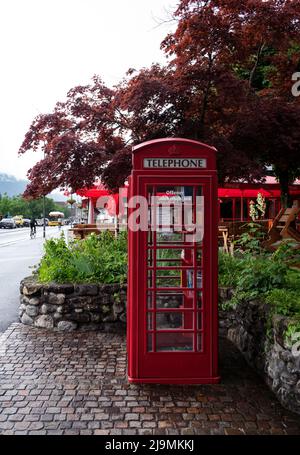 Blick auf ein öffentliches Swisscom-Telefon im klassischen britischen Stil in roter Telefonzelle mit einer offenen Bücherei in der Hauptstraße von Interlaken Stockfoto