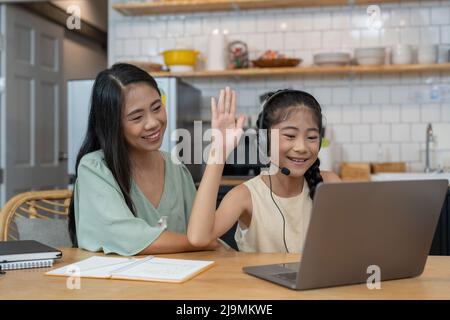 Glückliche asiatische Mutter und Kind sitzen mit Buntstiften am Küchentisch, nehmen per Videoanruf an einem virtuellen Zeichenkurs Teil, lächeln und winken hallo Stockfoto