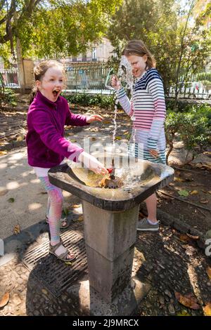 Junge Mädchen überraschten den hohen Druck eines Wasserbrunnens, als sie sich darauf vorbereitete, ein Getränk Wasser / Trinken aus einem durstlöschenden öffentlichen Brunnen draußen, in einem Stadtpark, in Catania, Sizilien, zu nehmen. Italien. (129) Stockfoto