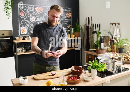 Porträt eines hübschen jungen erwachsenen kaukasischen Mannes mit Bart und Schnurrbart im Gesicht, der in der Küche steht und Zwiebel schneidet Stockfoto