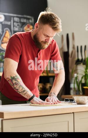 Bärtiger kaukasischer Mann in lässigem Outfit, der in der Loft-Küche steht und Teig für Pizza oder Kuchen knetet und abflacht Stockfoto