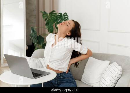 Eine junge Frau, die von zu Hause aus arbeitet, hat Rückenschmerzen aufgrund von Überlastung. Freiberuflerin knetet sich nach langer Arbeit auf der Couch den Rücken Stockfoto