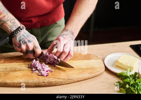 Nahaufnahme eines nicht erkennbaren Mannes, der mit einem Messer rote Zwiebeln auf einem Holzschneidebrett hackte Stockfoto