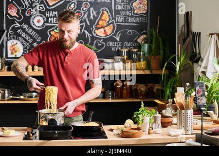 Porträt eines selbstbewussten bärtigen, reifen kaukasischen Influencers, der in der Loft-Küche steht und Pasta kocht Stockfoto