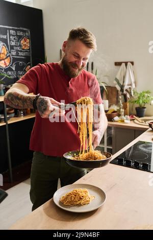 Fröhlicher kaukasischer Koch, der in der Loft-Küche steht und heiße Spaghetti mit Küchenzungen auf dem Teller serviert Stockfoto