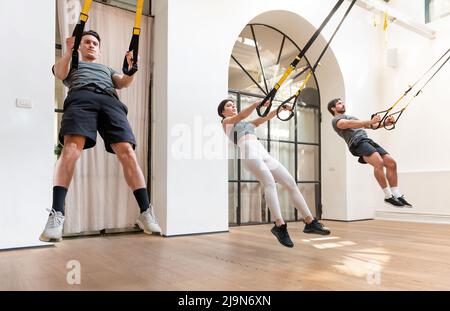 Gesellschaft von männlichen und weiblichen Athleten springen und hocken Übung mit TRX Seile während des aktiven Trainings im Fitnessstudio Stockfoto