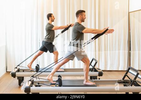 Seitenansicht Ganzkörper der athletischen Männer tun Baum Umarmungen Übung, während auf Reformern während Pilates-Klasse stehen Stockfoto