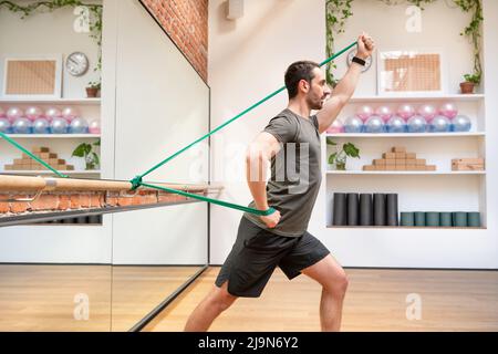 Seitenansicht des FIT-Männchens in der aktiven Trainingsbekleidung mit Gummiband, während er in der Nähe des Barres im Fitnessstudio steht Stockfoto