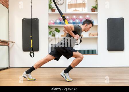 Seitenansicht des fokussierten Sportlers in Sportkleidung, der während des Suspensionstrainings im geräumigen Fitnessclub Übungen an TRX-Seilen macht Stockfoto