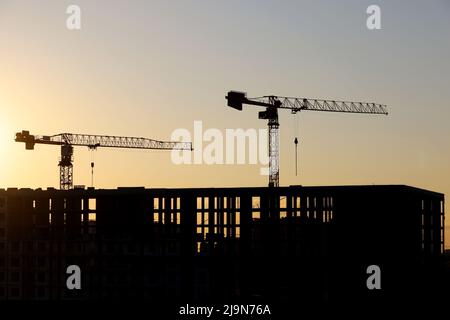 Silhouette von zwei Türmen Kran über unfertigen Gebäude bei Sonnenaufgang. Wohnungsbau, Wohnblock in der Stadt auf dem Hintergrund Stockfoto