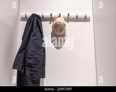 Blick aus der unteren Perspektive auf eine marineblaue Jacke und eine braune Baseballmütze, die in einem weißen, beleuchteten Schrank an einem Wandregal hing Stockfoto
