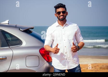 Indischer Geschäftsmann steht in der Nähe von Auto im Freien am Meeresstrand Sommer Guten Tag.ein Mann in einem weißen Hemd und schneeweißem Lächeln freut sich, ein neues Auto zu kaufen Stockfoto