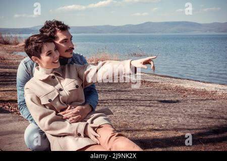 Schönes Paar am Ufer des Lake Bracciano sitzen. Das junge Paar umarmt sich beim Blick auf den Horizont. Platz für Ihre Nachricht kopieren. Stockfoto