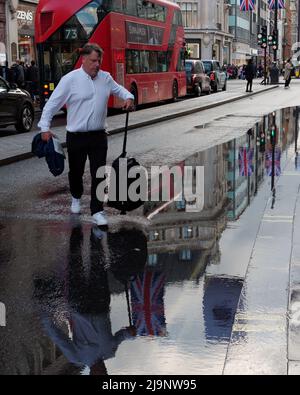 London, Greater London, England, Mai 11 2022: Männlicher Tourist trägt seinen Koffer über den Überschwemmungen in der Oxford Street nach heftigem Regen. Stockfoto