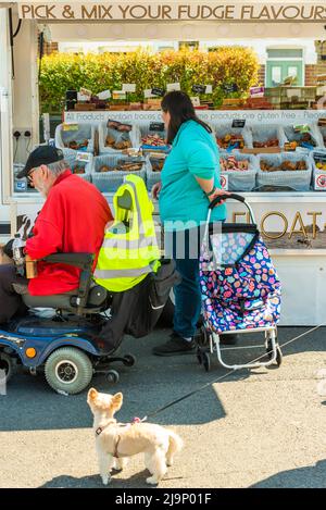 London, Penge, Vereinigtes Königreich - 30. April 2022: Maple Road Market Mann auf einem Mobilitätsroller und Frau mit einem Trolley entscheiden sich für Fudge Stockfoto