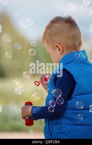 Ein kleiner Junge bläst Seifenblasen. Spiele mit Kindern in der Natur, an der frischen Luft im Sommer. Ein Kind spielt auf der Straße mit Seifenblasen. Stockfoto