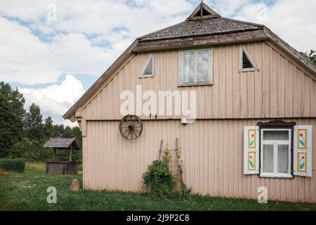 Altes traditionelles Holzhaus mit Fenster und Fensterläden, Rückansicht. Altes Kutschenhängerrad an der Wand, Holzbrunnen in der Nähe. Stockfoto