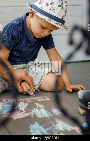 Der kleine Junge und seine Mutter zeichnen mit farbigen Kreiden auf einem Betonweg. Aktivitäten für Kinder im Freien im Sommer. Die Mutter steuert Stockfoto