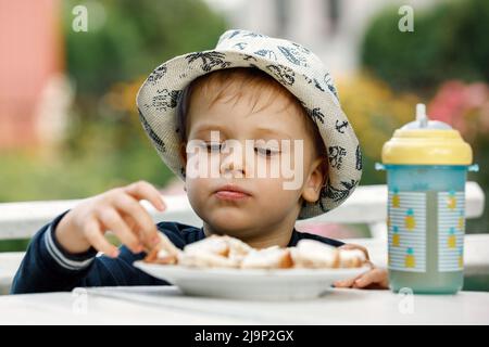 Der Junge isst Toast zum Frühstück mit Saft im Hintergrund eines Sommergartens. Ein Plastikbecher mit Ananassaft auf dem Tisch. Stockfoto