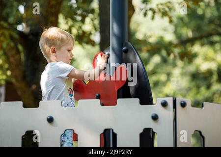 Kleiner Junge, der auf einem Spielplatz voller Zuversicht das Ruder eines Spielzeugschiffes dreht. Stockfoto