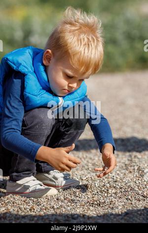 Ein kleiner Junge mit einer blauen Weste spielt draußen mit kleinen Kieselsteinen. Das Kind erkundet den Boden mit seinen eigenen Händen, er ist sehr an kleinen Kebb interessiert Stockfoto