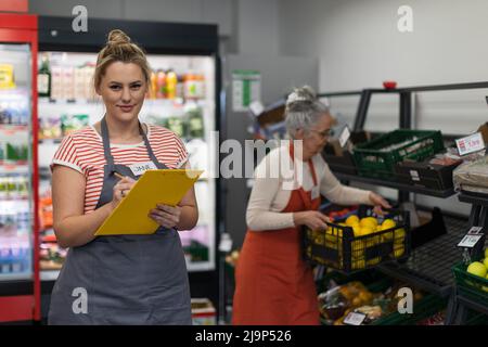 Junge Verkäuferin im Supermarkt in Gemüseschalen, im Background ist ihre Kollegin beim Befüllen von Vorräten. Stockfoto