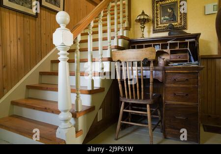 Antiker Schreibtisch mit Stuhl auf der Rückseite neben der Treppe, die zum Obergeschoss führt, in einem alten Haus im kanadischen Landhausstil aus dem Jahr 1741. Stockfoto