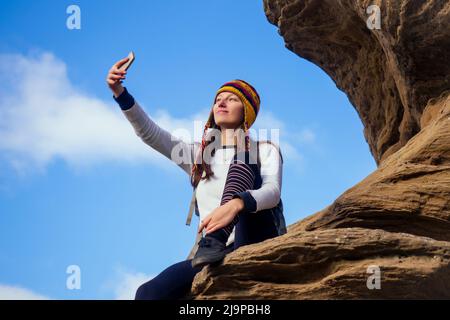 Schöne schlanke und sportliche junge Touristenfrau in einem lustigen Hut aus nepalesischer Wolle Yak sitzend und haben Ruhe machen Selfie-Handy Klettern großen Felsen klettert auf Stockfoto