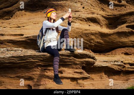 Schöne schlanke und sportliche junge Touristenfrau in einem lustigen Hut aus nepalesischer Wolle Yak sitzend und haben Ruhe machen Selfie-Handy Klettern großen Felsen klettert auf Stockfoto